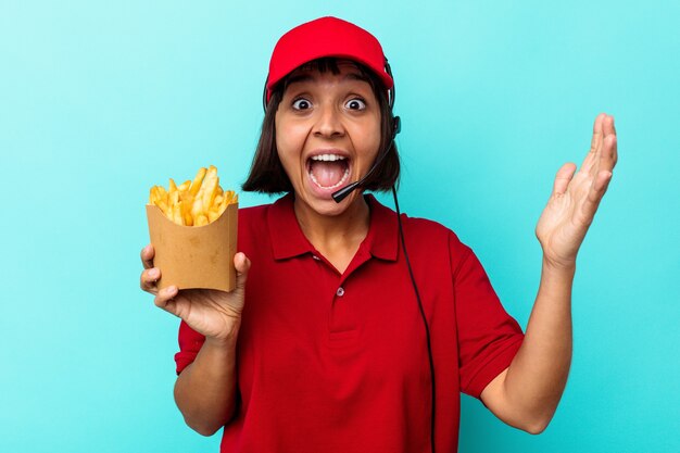 Young mixed race woman fast food restaurant worker holding fries isolated on blue background receiving a pleasant surprise, excited and raising hands.