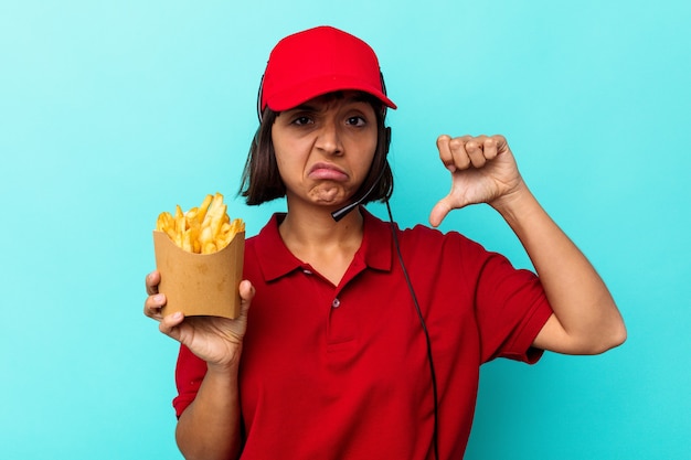 Young mixed race woman fast food restaurant worker holding fries isolated on blue background showing a dislike gesture, thumbs down. Disagreement concept.