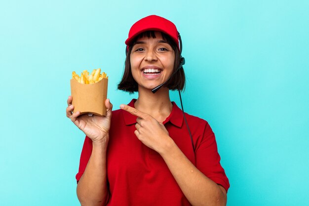 Young mixed race woman fast food restaurant worker holding fries isolated on blue background smiling and pointing aside, showing something at blank space.