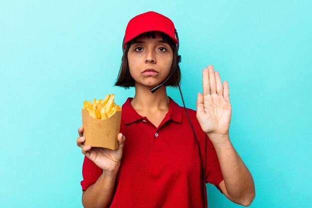 Young mixed race woman fast food restaurant worker holding fries isolated on blue background standing with outstretched hand showing stop sign, preventing you.