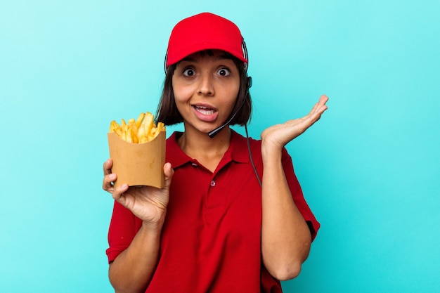 Young mixed race woman fast food restaurant worker holding fries isolated on blue background surprised and shocked.