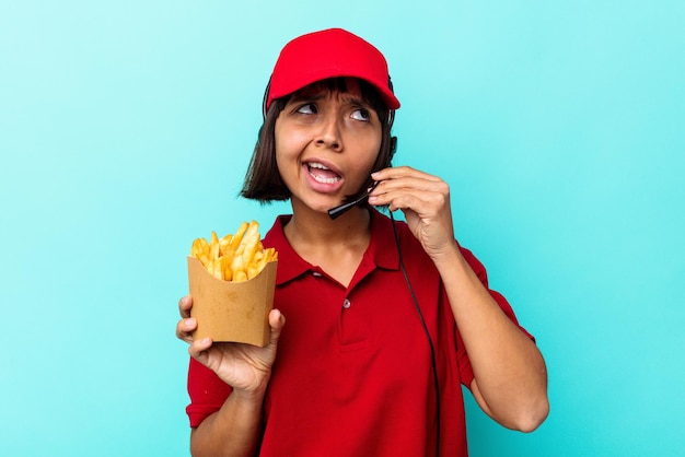 Young mixed race woman fast food restaurant worker holding fries isolated on blue background trying to listening a gossip.