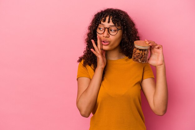 Young mixed race woman holding an almond jar isolated on pink background is saying a secret hot braking news and looking aside