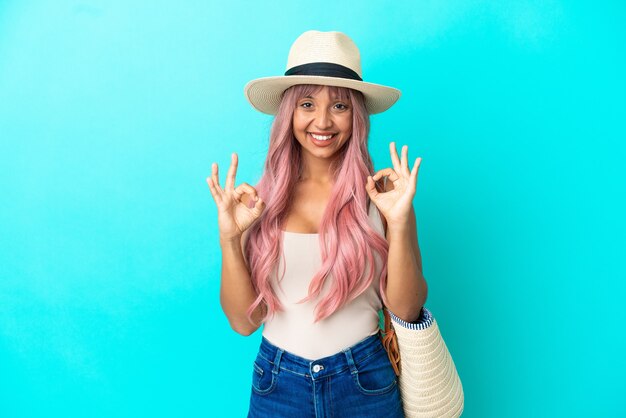 Young mixed race woman holding a beach bag with pamela isolated on blue background showing an ok sign with fingers