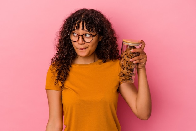 Young mixed race woman holding a cookies jar isolated on pink background confused, feels doubtful and unsure.