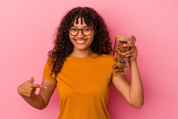 Young mixed race woman holding a cookies jar isolated on pink background person pointing by hand to a shirt copy space, proud and confident