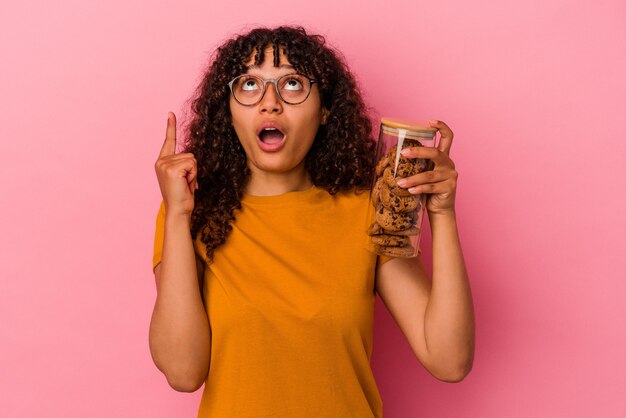 Young mixed race woman holding a cookies jar isolated on pink background pointing upside with opened mouth.