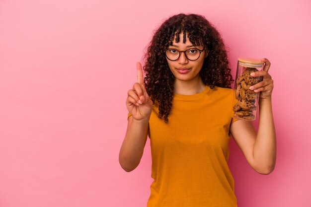 Young mixed race woman holding a cookies jar isolated on pink background showing number one with finger.