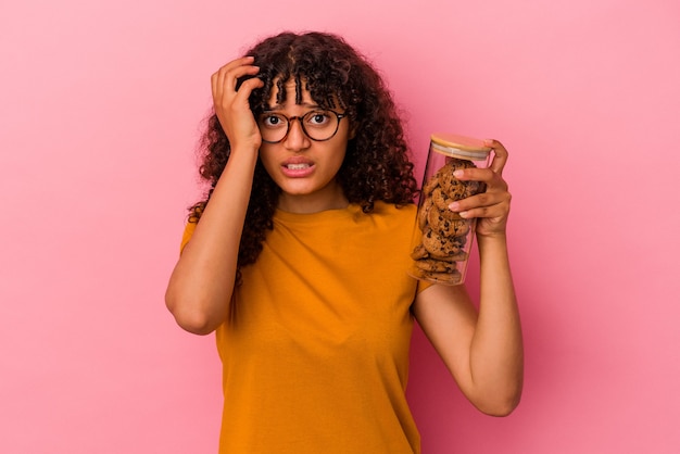 Young mixed race woman holding a cookies jar isolated on pink wall being shocked, she has remembered important meeting.