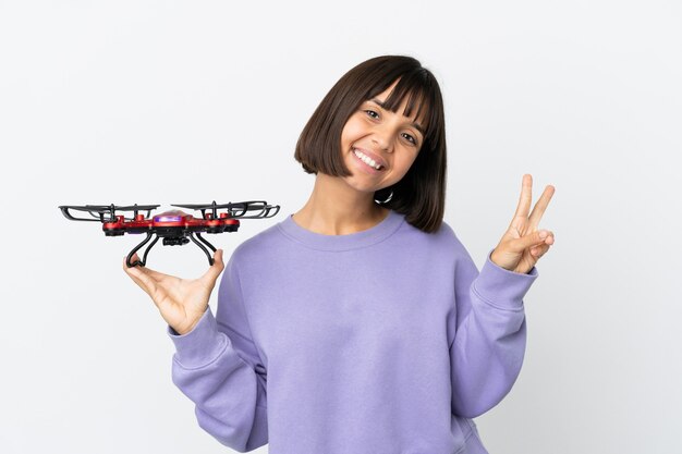 Young mixed race woman holding a drone isolated on white background smiling and showing victory sign