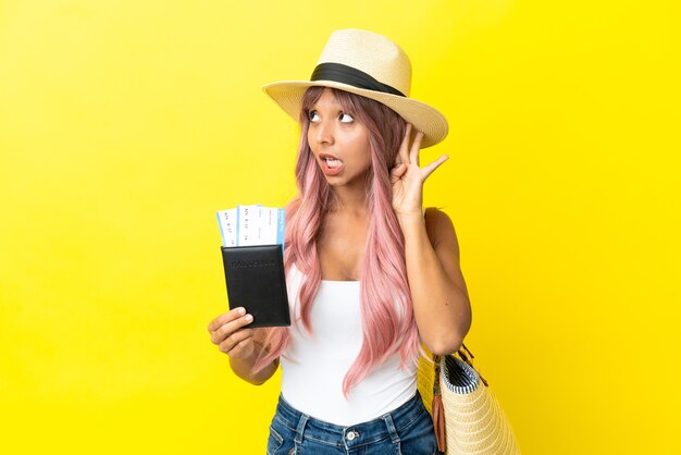 Young mixed race woman holding passport and beach bag isolated on yellow background listening to something by putting hand on the ear