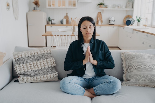 Young mixed race woman is practicing yoga sitting on couch in lotus pose with her eyes closed