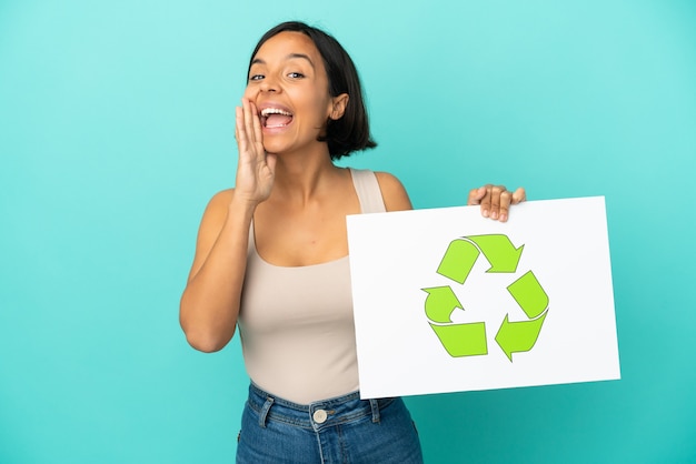 Young mixed race woman isolated on blue background holding a placard with recycle icon and shouting