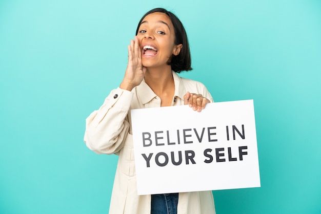 Young mixed race woman isolated on blue background holding a placard with text Believe In Your Self and shouting