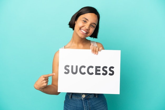 Young mixed race woman isolated on blue background holding a placard with text SUCCESS and  pointing it