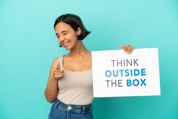 Young mixed race woman isolated on blue background holding a placard with text Think Outside The Box and pointing to the front