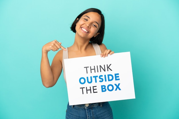 Young mixed race woman isolated on blue background holding a placard with text Think Outside The Box and  pointing it