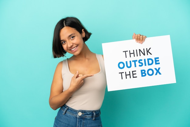 Young mixed race woman isolated on blue background holding a placard with text Think Outside The Box and  pointing it