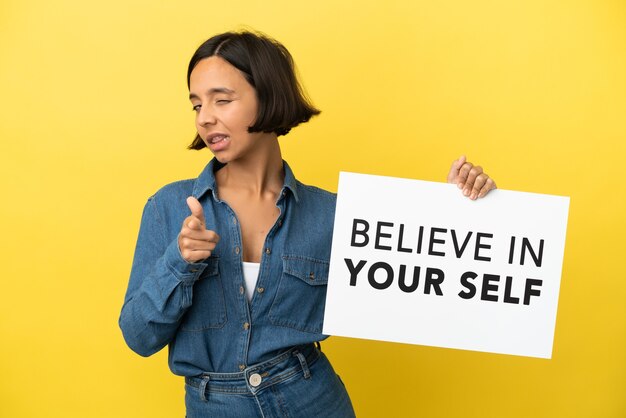 Young mixed race woman isolated on yellow background holding a placard with text Believe In Your Self and pointing to the front