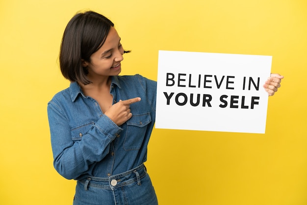 Young mixed race woman isolated on yellow background holding a placard with text Believe In Your Self and pointing it