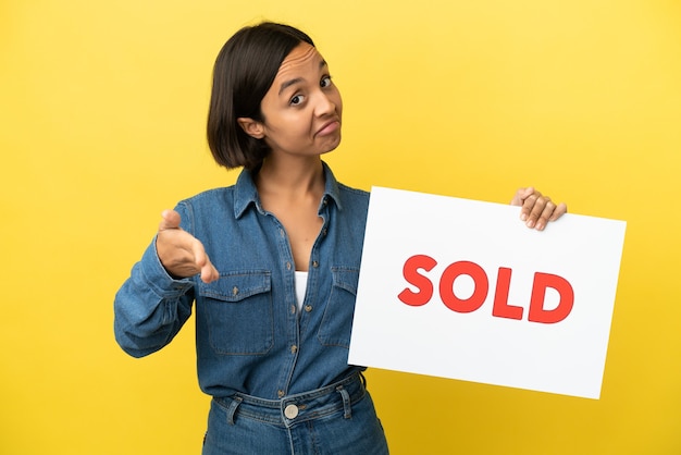 Young mixed race woman isolated on yellow background holding a placard with text SOLD making a deal