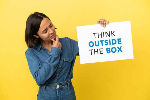 Young mixed race woman isolated on yellow background holding a placard with text Think Outside The Box and thinking