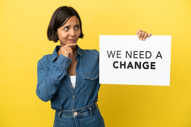 Young mixed race woman isolated on yellow background holding a placard with text We Need a Change and thinking