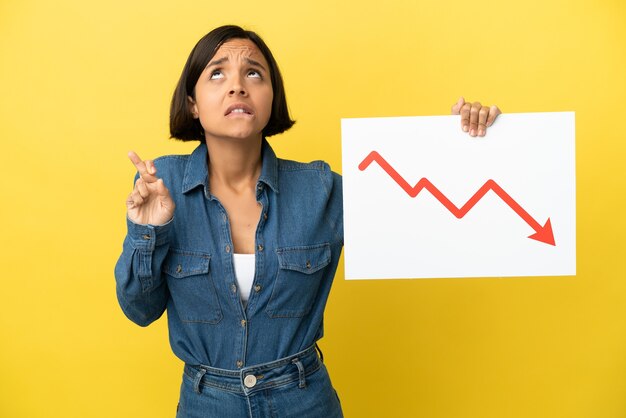 Young mixed race woman isolated on yellow background holding a sign with a decreasing statistics arrow symbol with fingers crossing