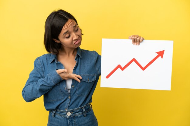 Young mixed race woman isolated on yellow background holding a sign with a growing statistics arrow symbol and  pointing it