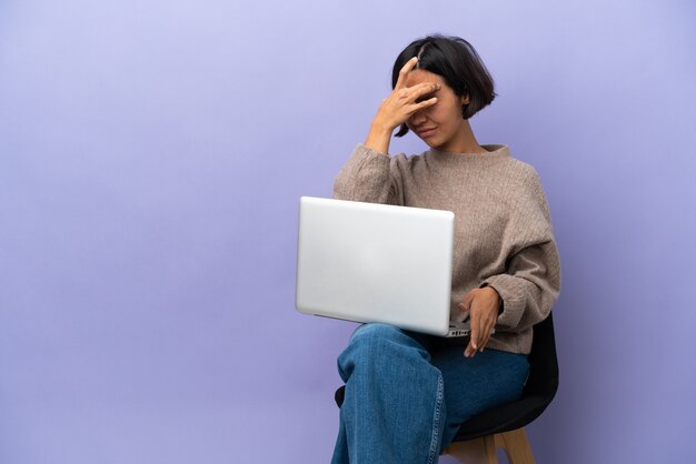 Young mixed race woman sitting on a chair with laptop isolated on purple background with headache