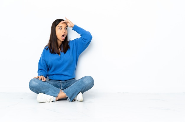 Young mixed race woman sitting on the floor isolated on white background doing surprise gesture while looking to the side