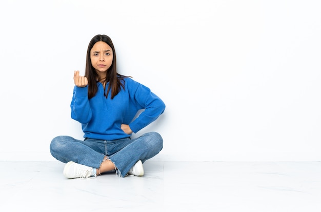 Young mixed race woman sitting on the floor isolated on white background making Italian gesture