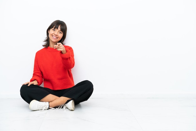 Young mixed race woman sitting on the floor isolated on white background points finger at you with a confident expression