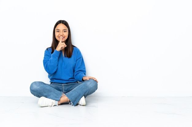 Young mixed race woman sitting on the floor isolated on white background showing a sign of silence gesture putting finger in mouth