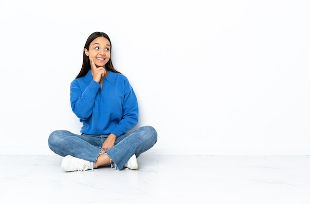 Young mixed race woman sitting on the floor isolated on white background thinking an idea while looking up