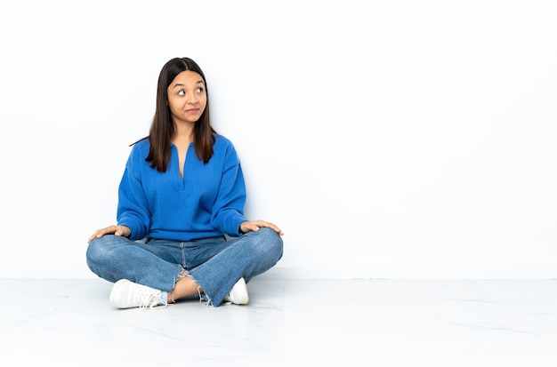 Young mixed race woman sitting on the floor isolated on white  making doubts gesture looking side