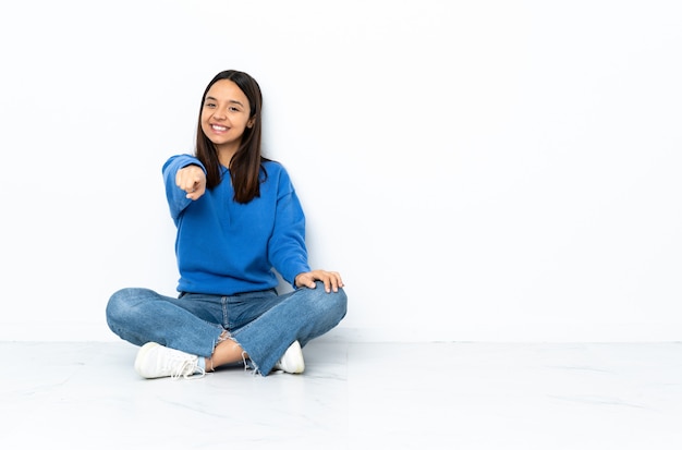 Young mixed race woman sitting on the floor isolated on white pointing front with happy expression