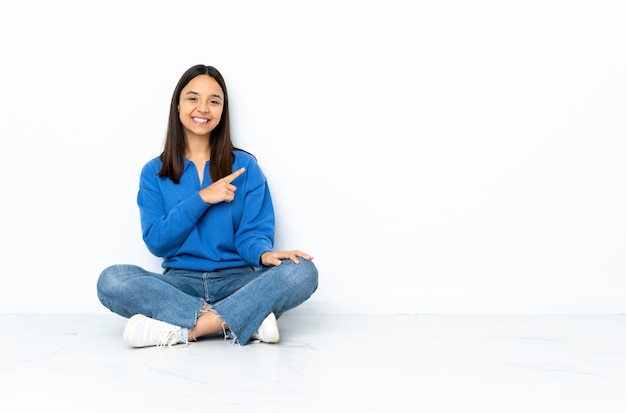 Young mixed race woman sitting on the floor isolated on white pointing to the side to present a product