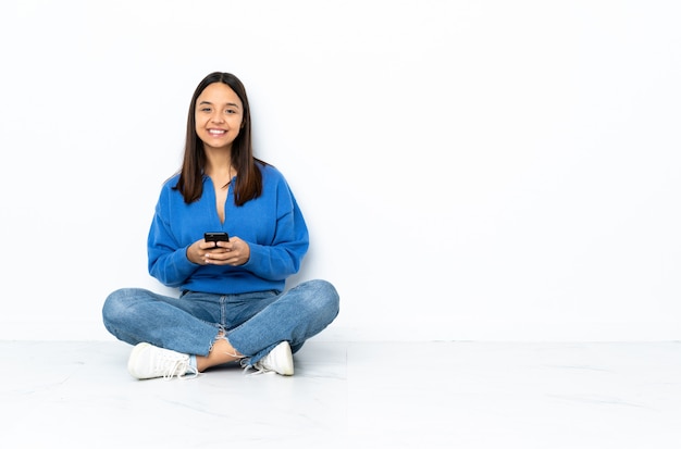 Young mixed race woman sitting on the floor isolated on white sending a message with the mobile