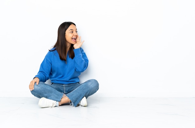 Young mixed race woman sitting on the floor isolated on white shouting with mouth wide open to the side