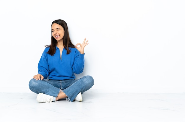 Young mixed race woman sitting on the floor isolated on white  showing ok sign with fingers