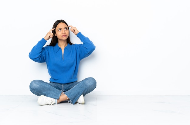 Young mixed race woman sitting on the floor isolated on white wall having doubts and thinking