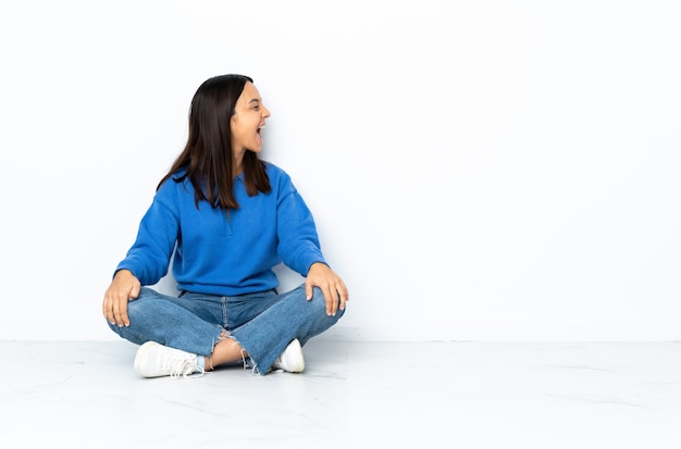 Young mixed race woman sitting on the floor isolated on white wall laughing in lateral position