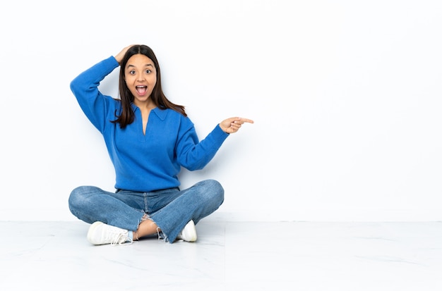 Young mixed race woman sitting on the floor isolated on white wall surprised and pointing finger to the side