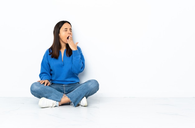 Young mixed race woman sitting on the floor isolated on white yawning and covering wide open mouth with hand