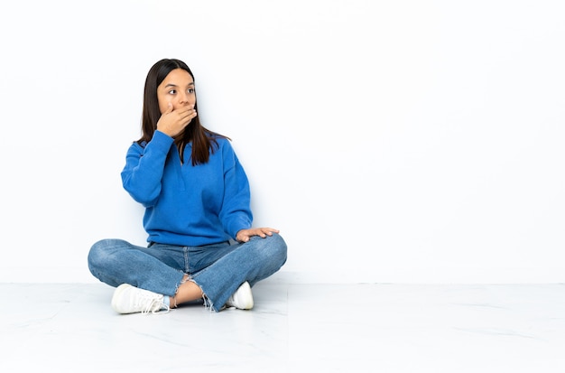 Young mixed race woman sitting on the floor on white wall doing surprised gesture while looking to the side