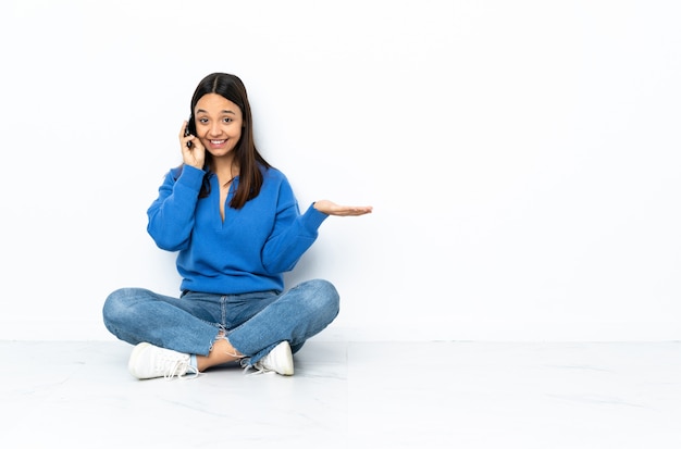 Young mixed race woman sitting on the floor on white wall keeping a conversation with the mobile phone with someone