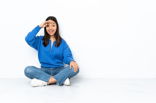 Young mixed race woman sitting on the floor on white wall saluting with hand with happy expression