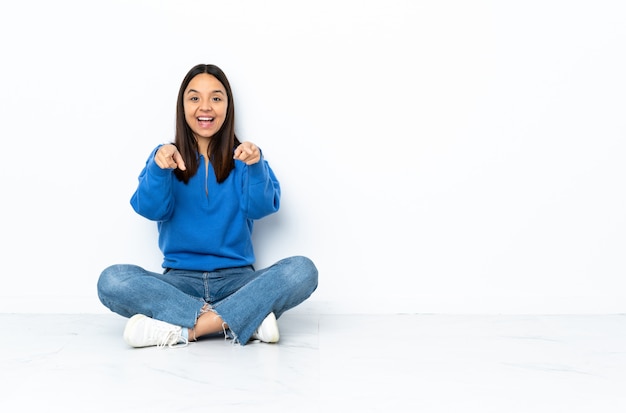 Young mixed race woman sitting on the floor on white wall surprised and pointing front