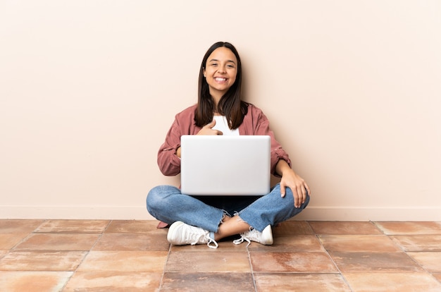 Young mixed race woman with a laptop sitting on the floor giving a thumbs up gesture